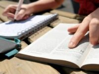 Mão escrevendo em caderno analisando Bíblia